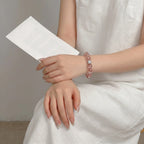 Frontal mid-shot of a woman with crossed legs resting a hand on her knee, with a paper over the left wrist showing a crystal bracelet.