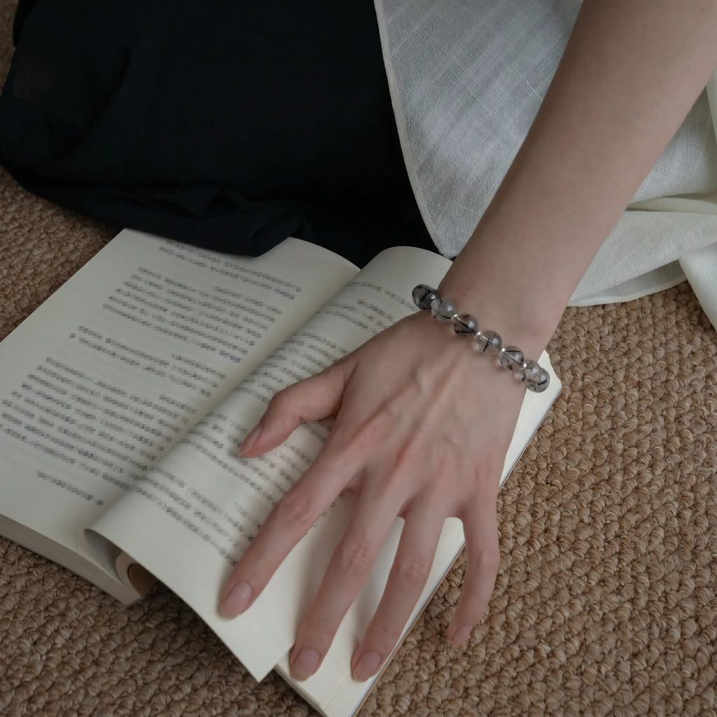 Left-side detail shot of a female hand pressing down on a book placed flat on the floor revealing the gemstone bracelet.