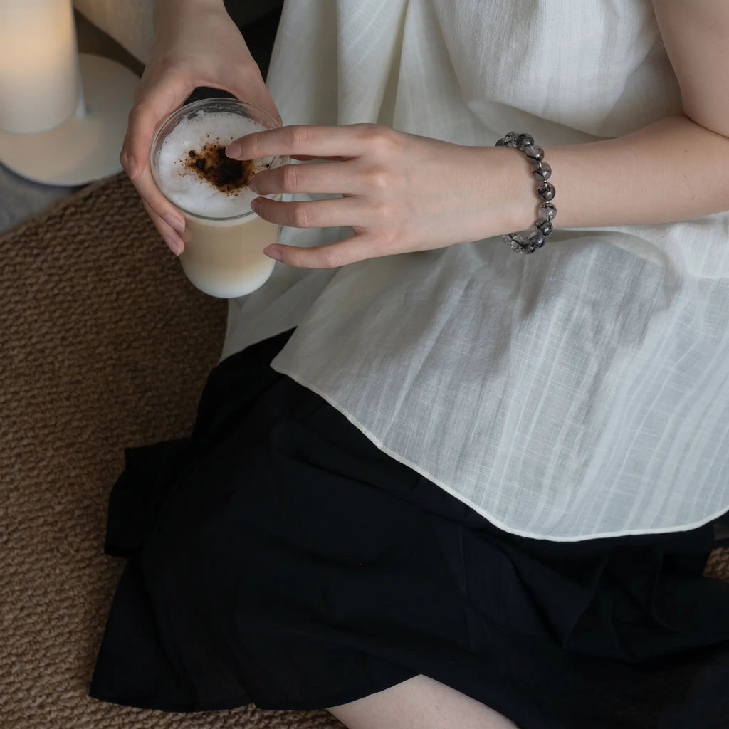 Top-down high angle shot from the left side showing a woman by the bed holding a coffee cup with a crystal needle quartz bracelet.