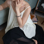 Left-side perspective of a woman sitting on the floor leaning against a bed, touching her neck to show the black crystal bracelet.