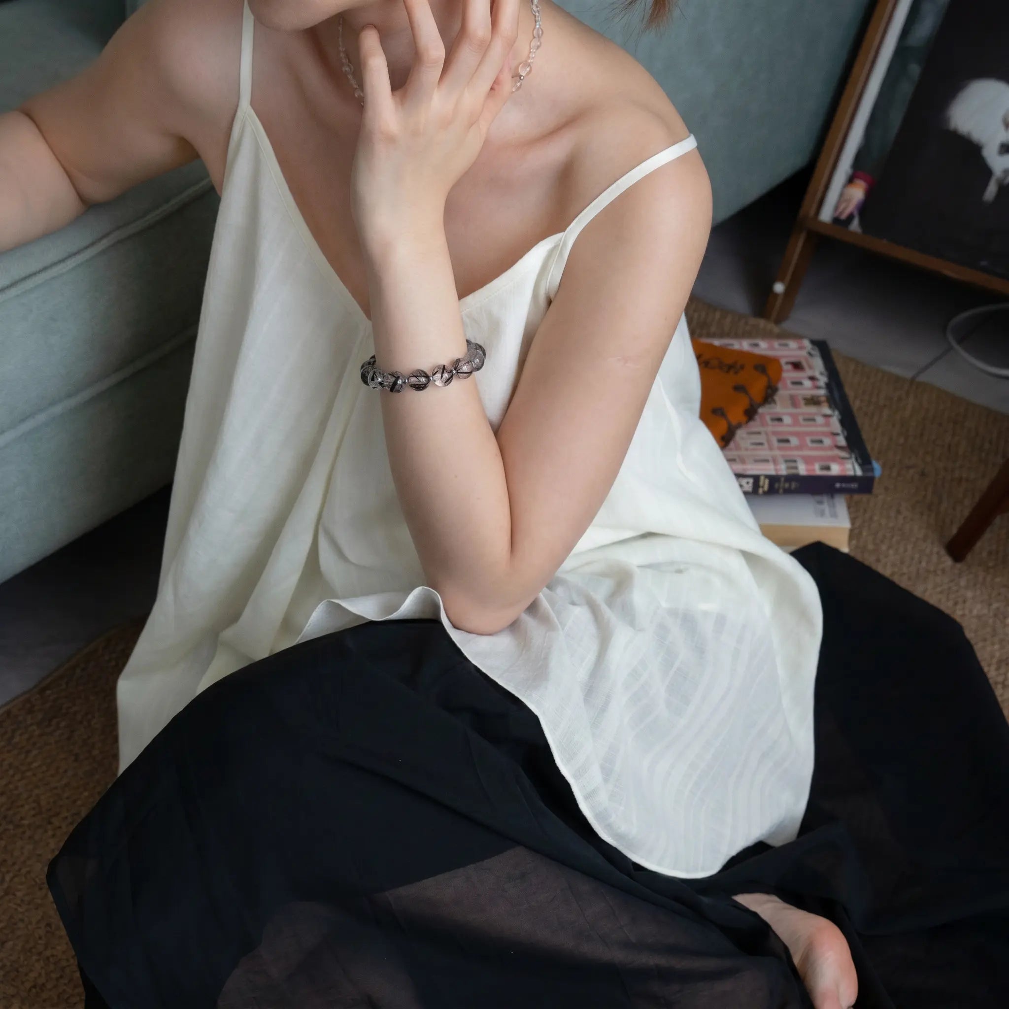 Left-side perspective of a woman sitting on the floor leaning against a bed, touching her neck to show the black crystal bracelet.