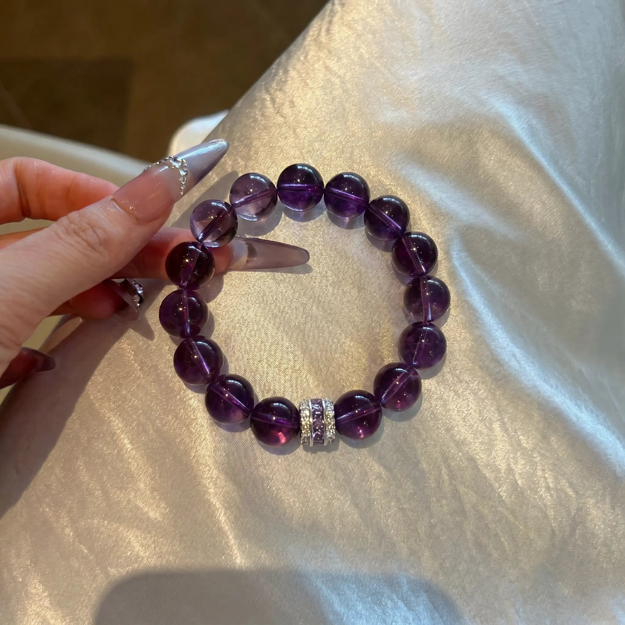 Close-up shot of a hand holding the Eternal Elegant Amethyst Bracelet resting gracefully over shining white silk fabric.