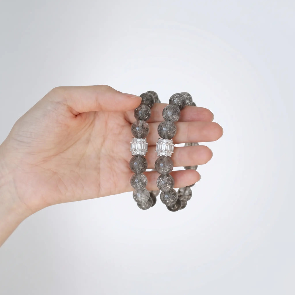 Close-up shot of a single left hand holding two layered clear crystal quartz bracelets against a plain white backdrop.