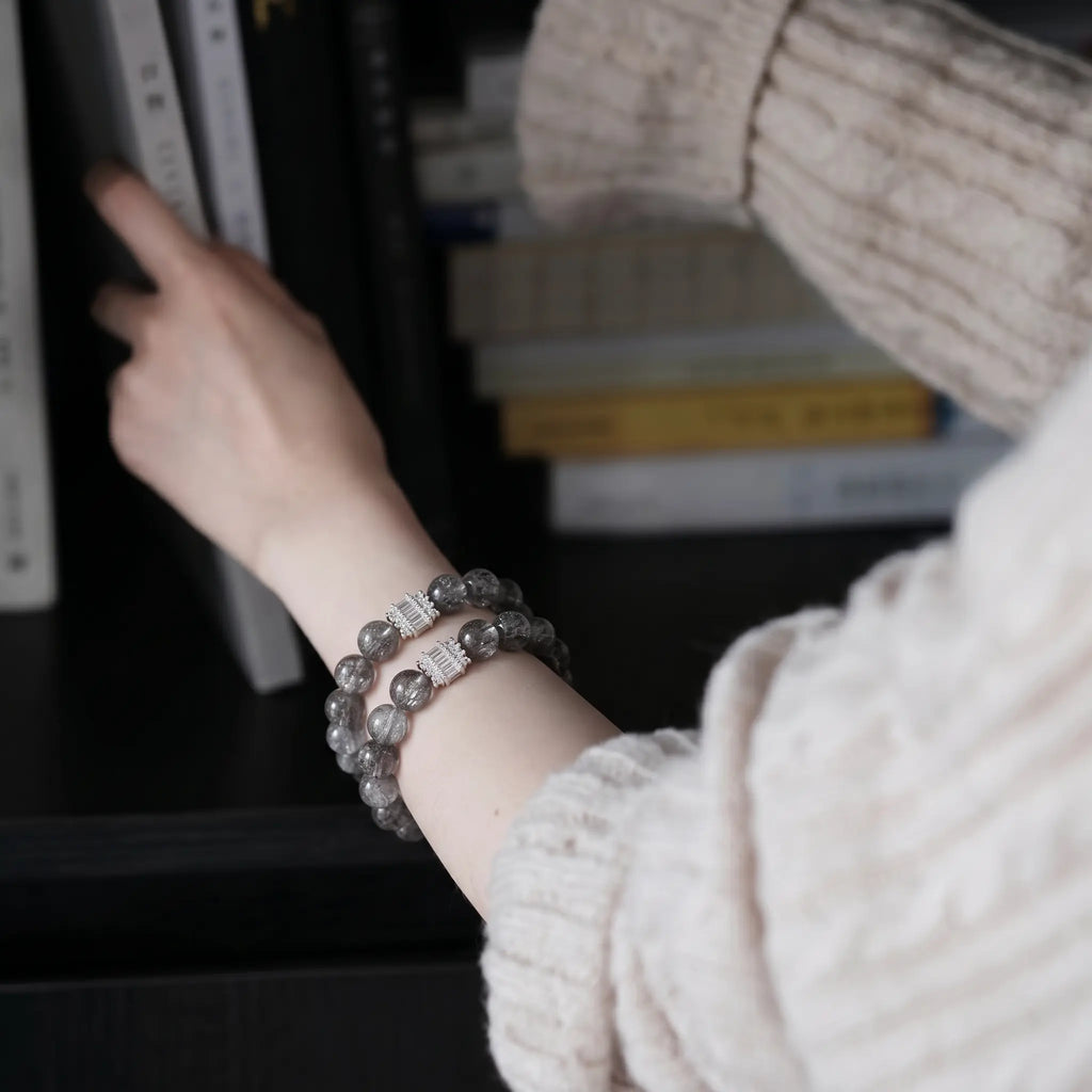 Over-the-shoulder perspective of a woman hand browsing books on a wooden bookshelf displaying her crystal bracelet.