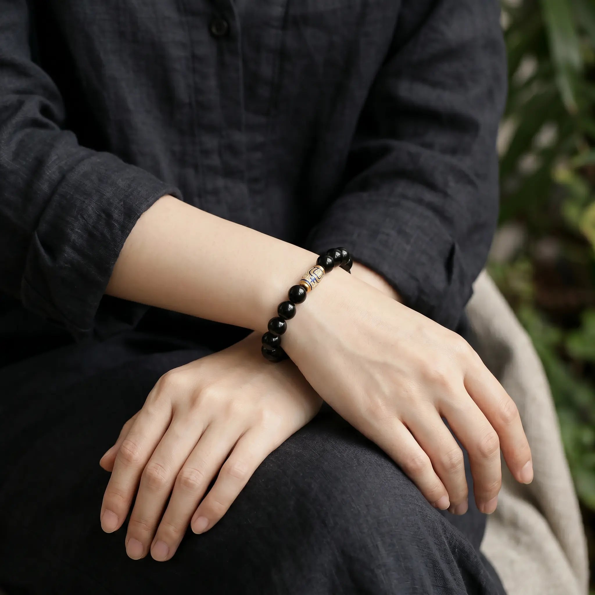 Detailed view of a woman with crossed legs and hands folded over knees revealing an artisan enameled obsidian bracelet.