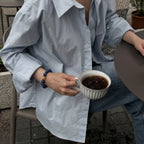 Side perspective view of a woman seated in a chair holding a porcelain coffee cup with her wrist decorated with a mineral gemstone bracelet.