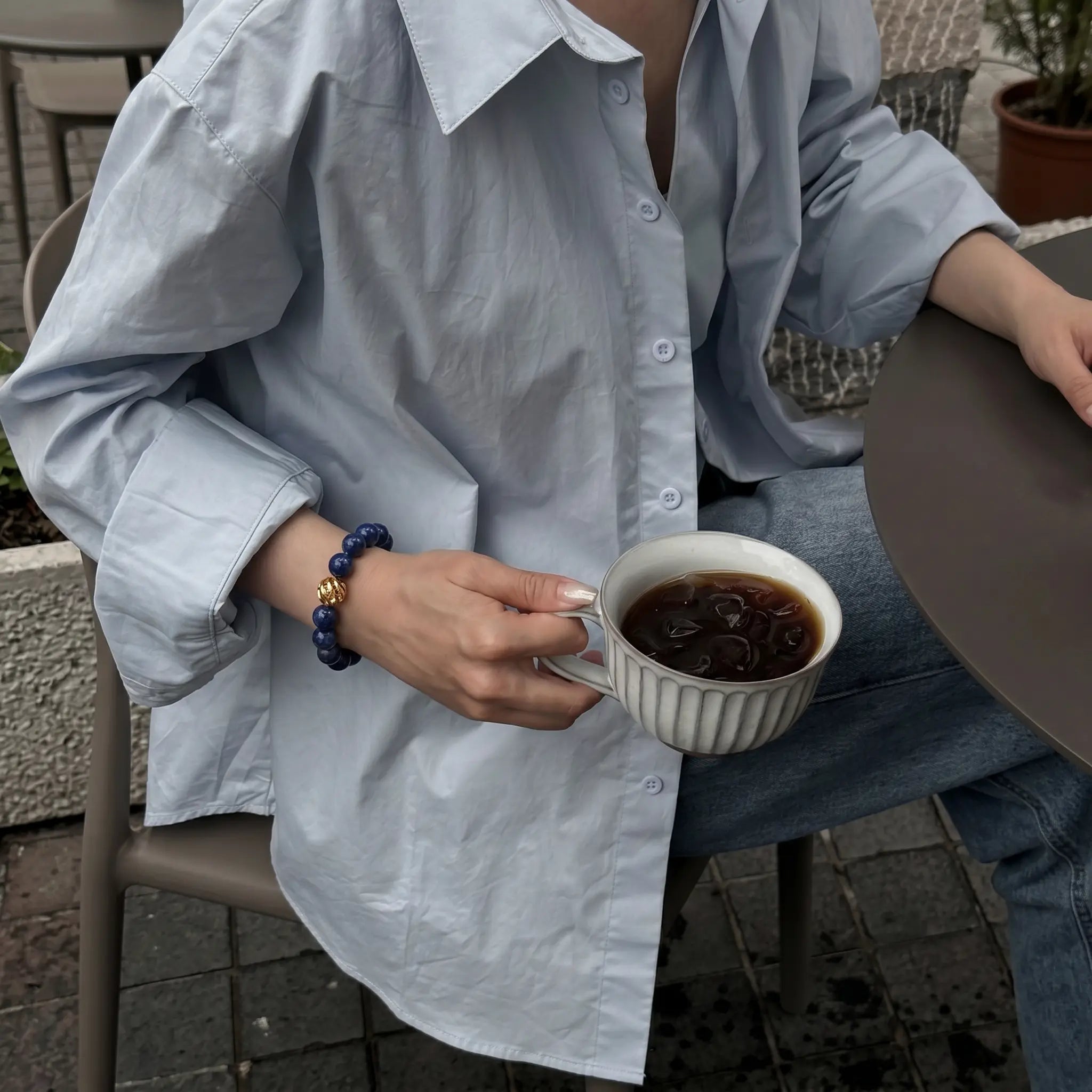 Side perspective view of a woman seated in a chair holding a porcelain coffee cup with her wrist decorated with a mineral gemstone bracelet.