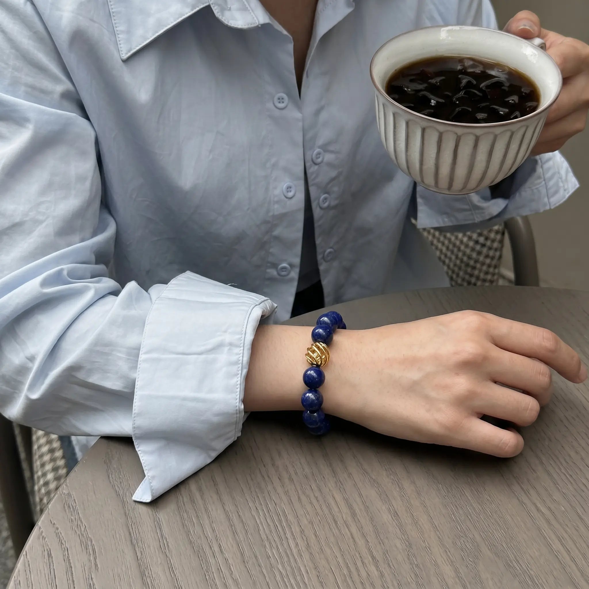 Top-down view of a woman seated at a desk with her right wrist adorned with a gemstone bracelet resting flat, while her left hand lifts a coffee cup.