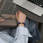 Over-the-shoulder view focusing on a woman right hand resting over a luxury handbag placed on her lap, showing the crystal bracelet.