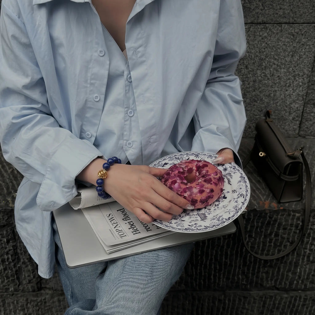 High angle view of a woman with crossed legs and a laptop plus newspaper on her lap, her right arm reaching for a donut on a plate.