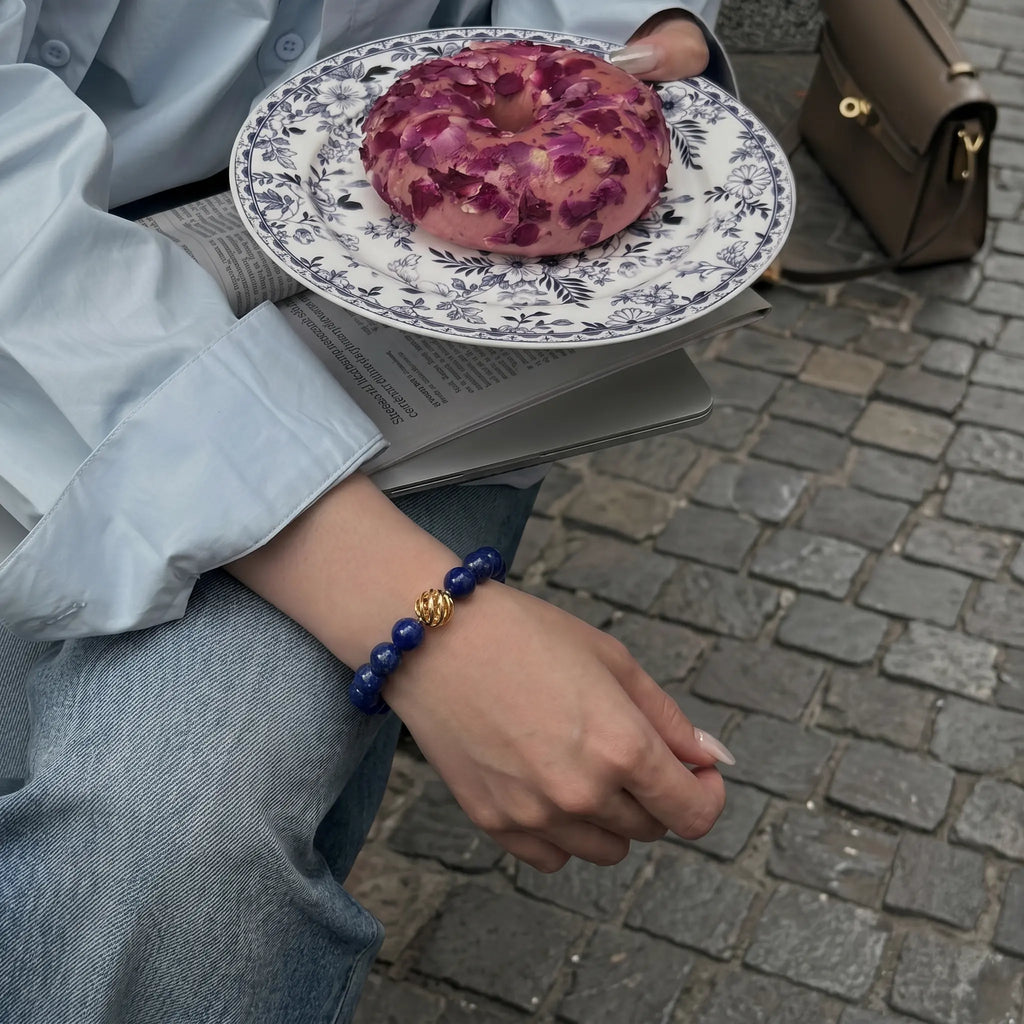 Close-up shot of a woman with crossed legs resting her right arm downward while her left hand holds a sweet donut, revealing a gold sheen lapis bracelet.