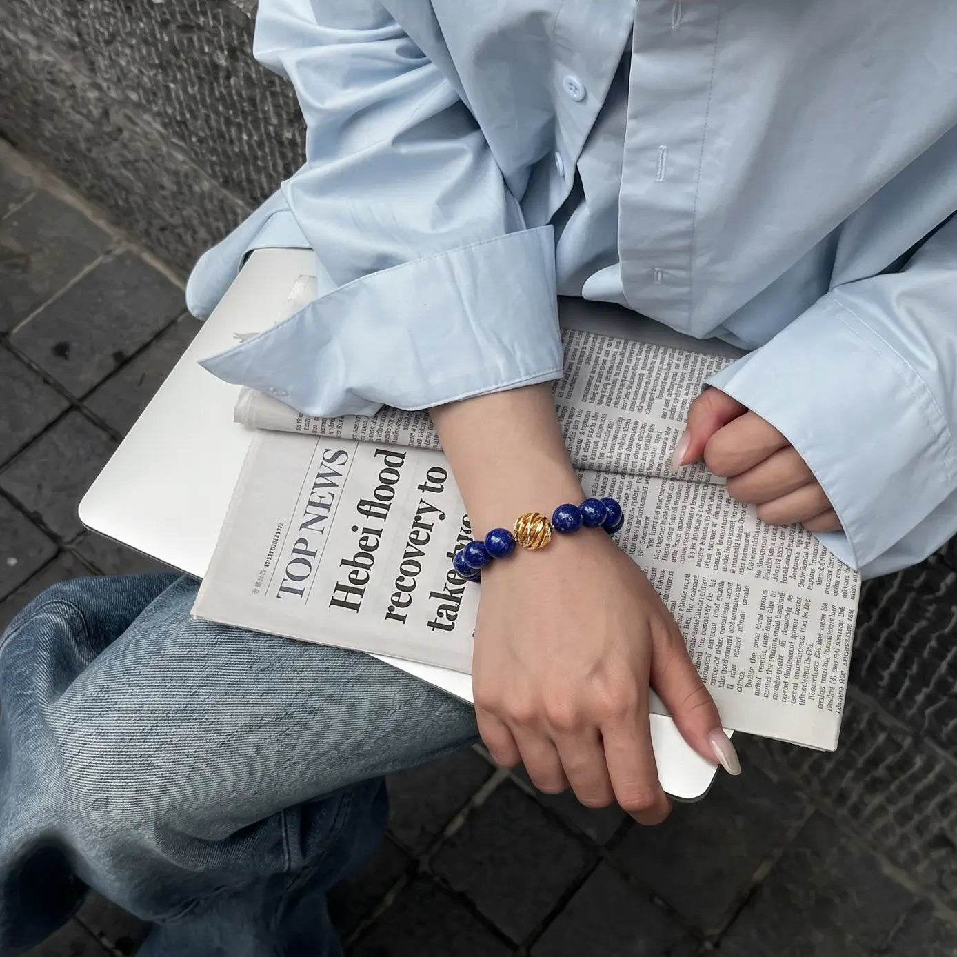 High angle diagonal shot of a sitting woman with her right hand resting over a book and laptop on her lap, showing a lapis lazuli bracelet.