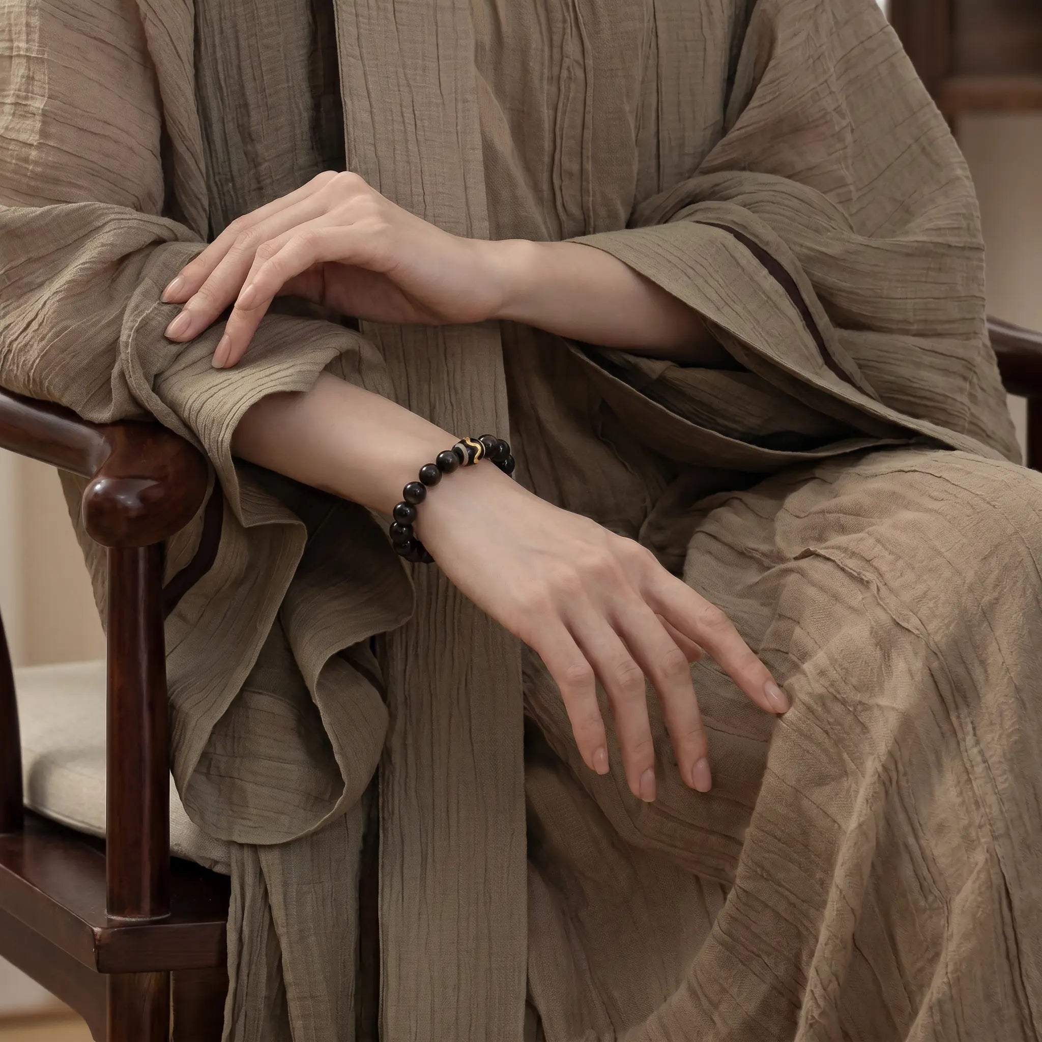 Detailed view of an elegant lady seated on a wooden chair with her right elbow on the armrest revealing a black gemstone bracelet.