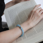 First-person POV view of a woman open palm upward displaying the ocean s grace aquamarine bracelet over open printed book pages.