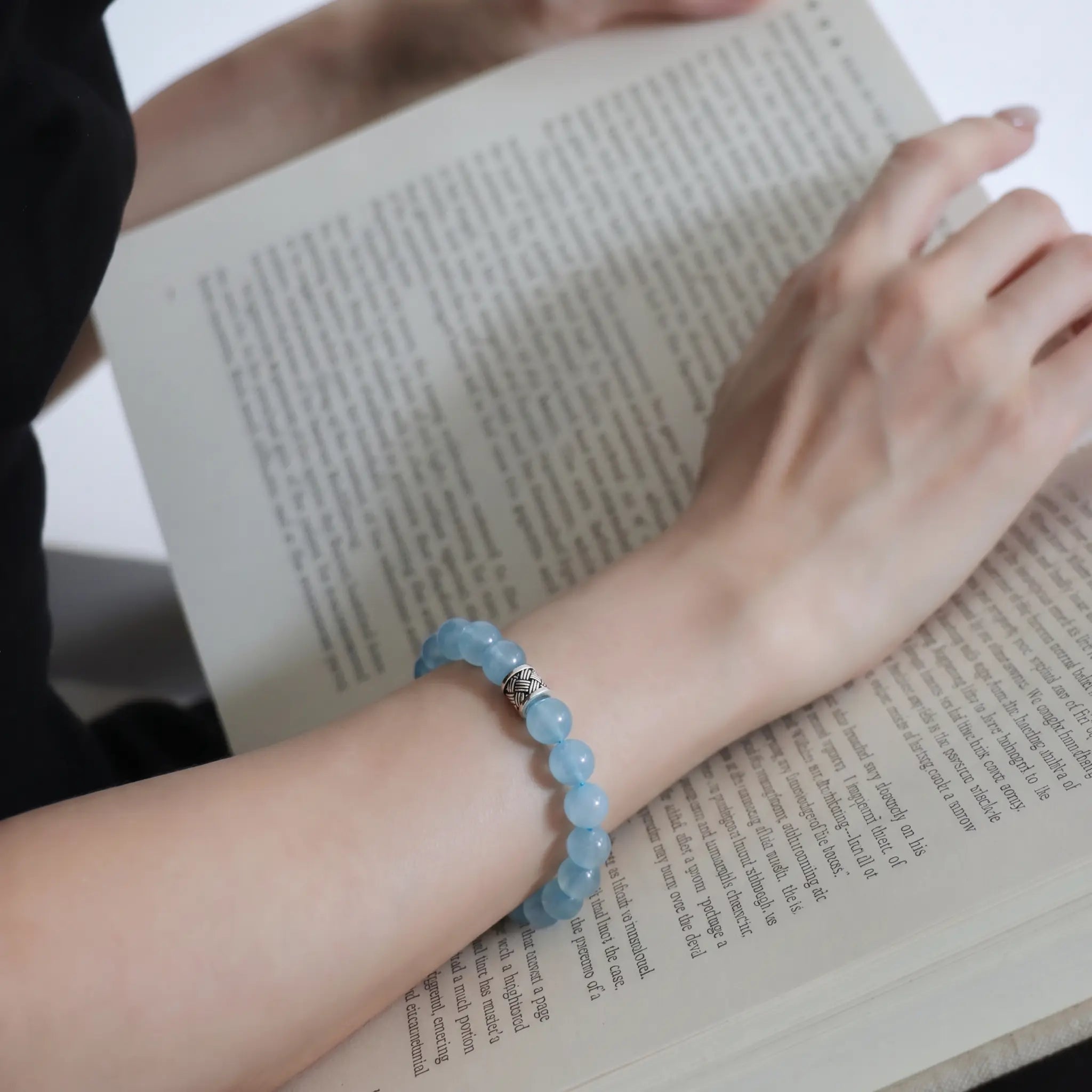 First-person POV view of a woman open palm upward displaying the ocean s grace aquamarine bracelet over open printed book pages.