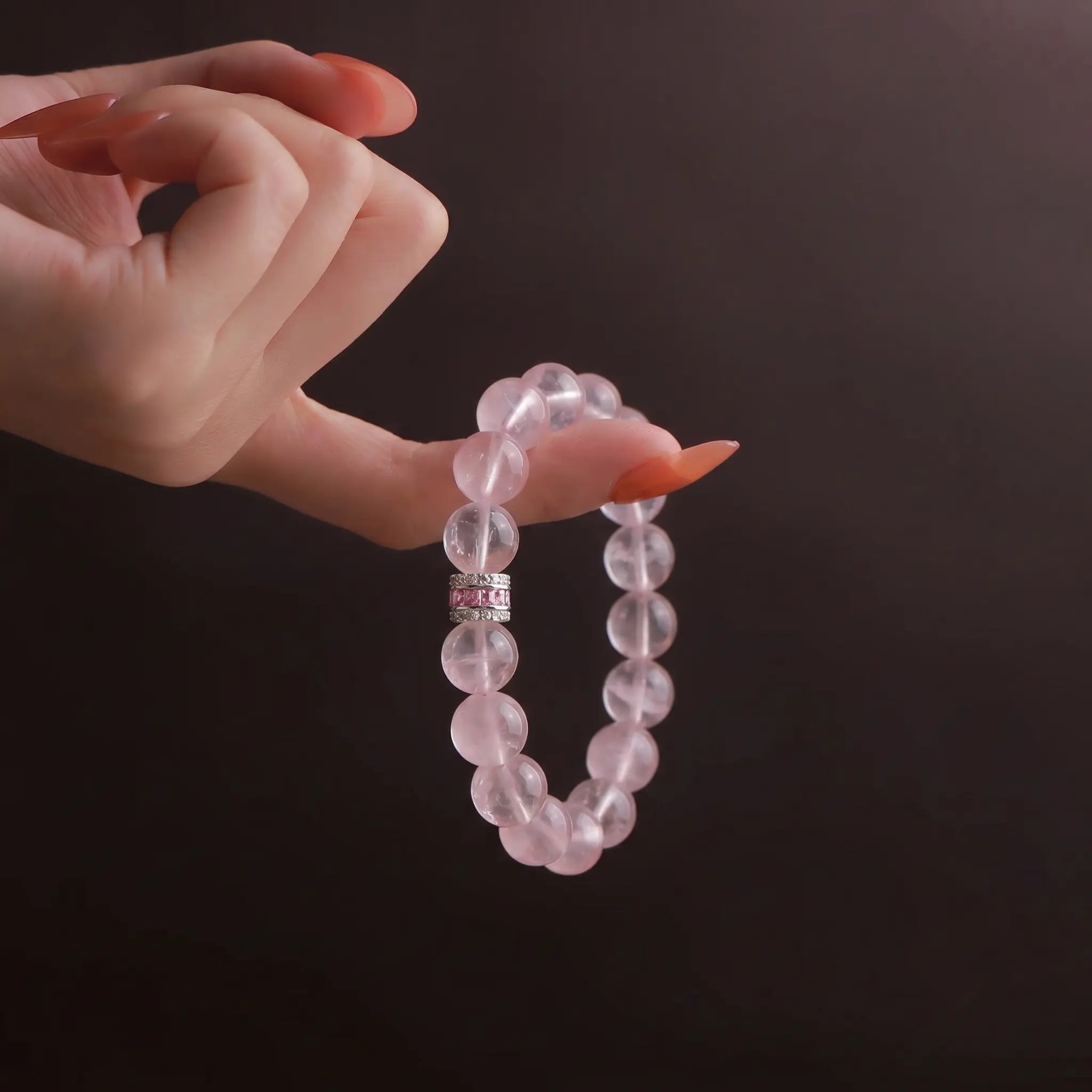 Detailed look of Pure Heat rose quartz bracelet hanging elegantly from a single finger against a dark background.