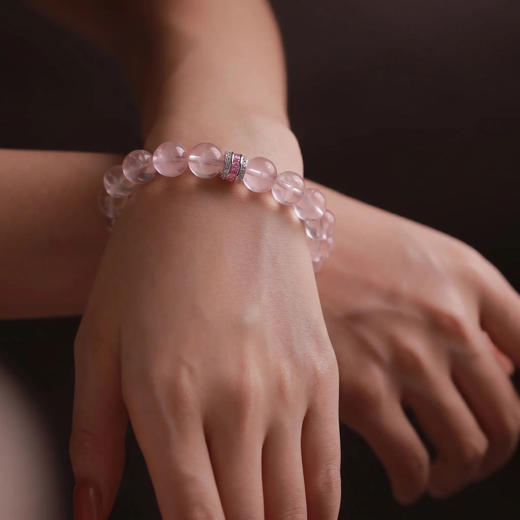 Close-up shot of overlapping hands gracefully displaying the Pure Heat rose quartz bracelet against a muted dark background.