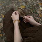 Over-the-shoulder macro perspective of hands resting on legs holding a leaf revealing the crystal quartz bracelet on the wrist.