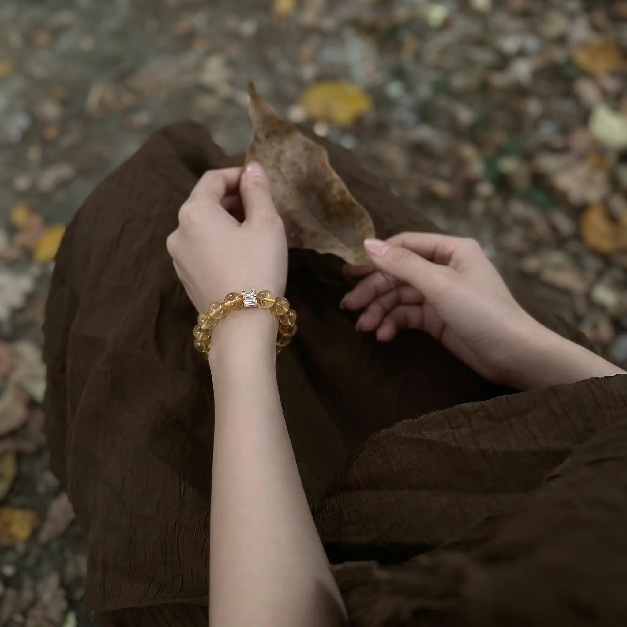 Over-the-shoulder macro perspective of hands resting on legs holding a leaf revealing the crystal quartz bracelet on the wrist.