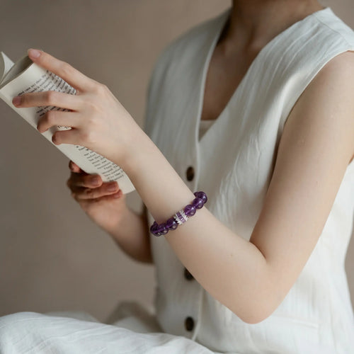 Left-side view of an elegant woman seated comfortably holding a book with a purple crystal bracelet on her left wrist.