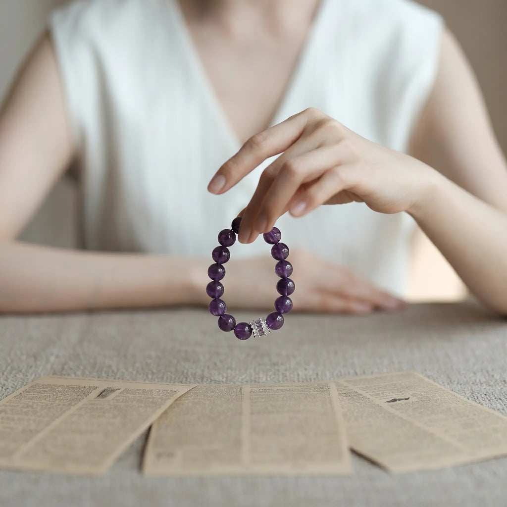Front view of a woman at a desk with three sheets of paper, dangling a purple gemstone bracelet from her fingers in a mystical gesture.