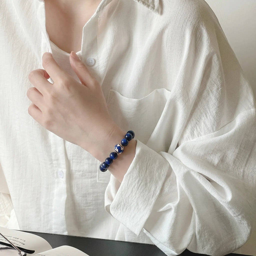 Side portrait view of a woman with her elbow resting on a desk gently touching her collar to reveal a deep blue lapis lazuli bracelet.