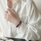 Side portrait view of a woman with her elbow resting on a desk gently touching her collar to reveal a deep blue lapis lazuli bracelet.