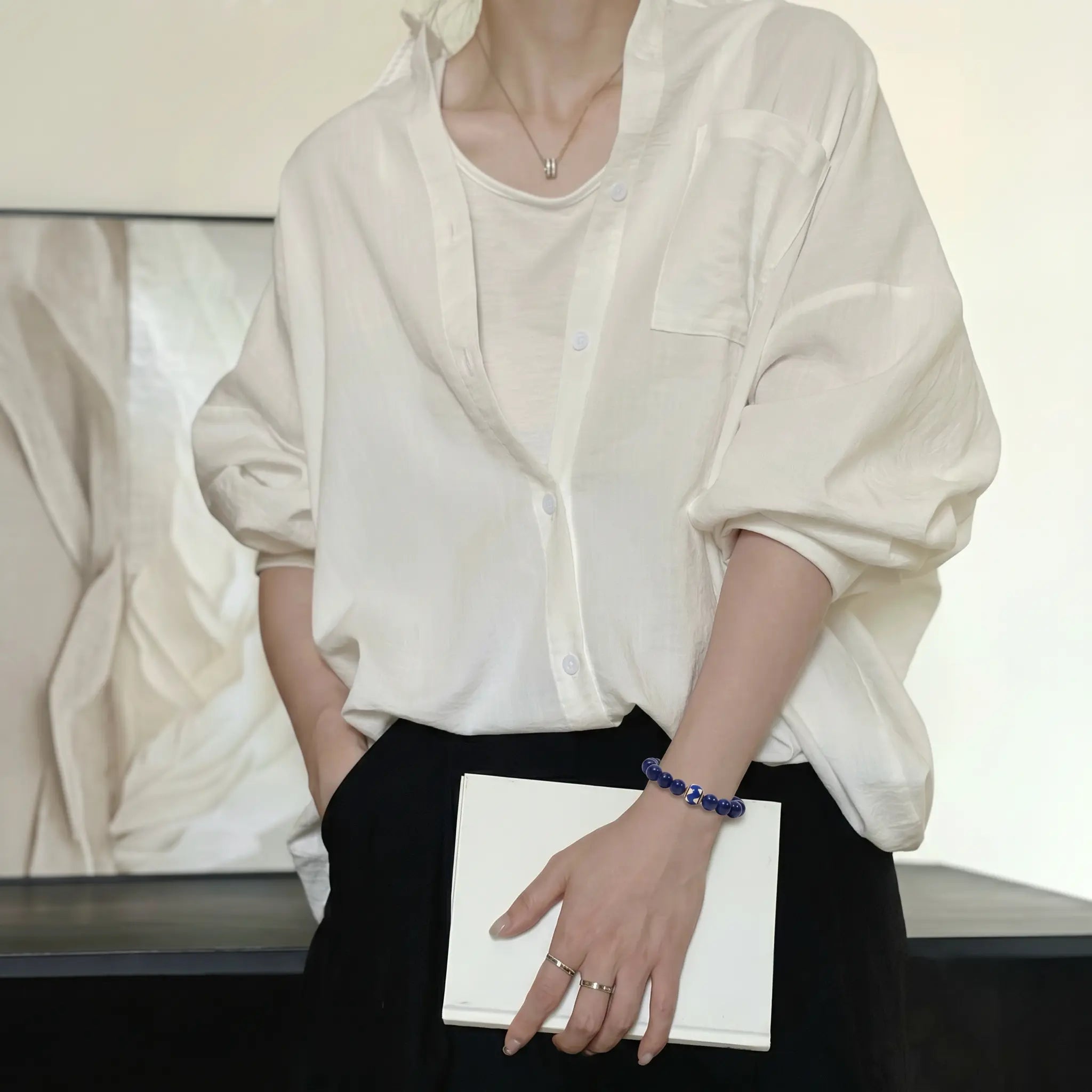 Mid-shot portrait of a woman holding a book over her dress while her right hand is tucked into a pants pocket.