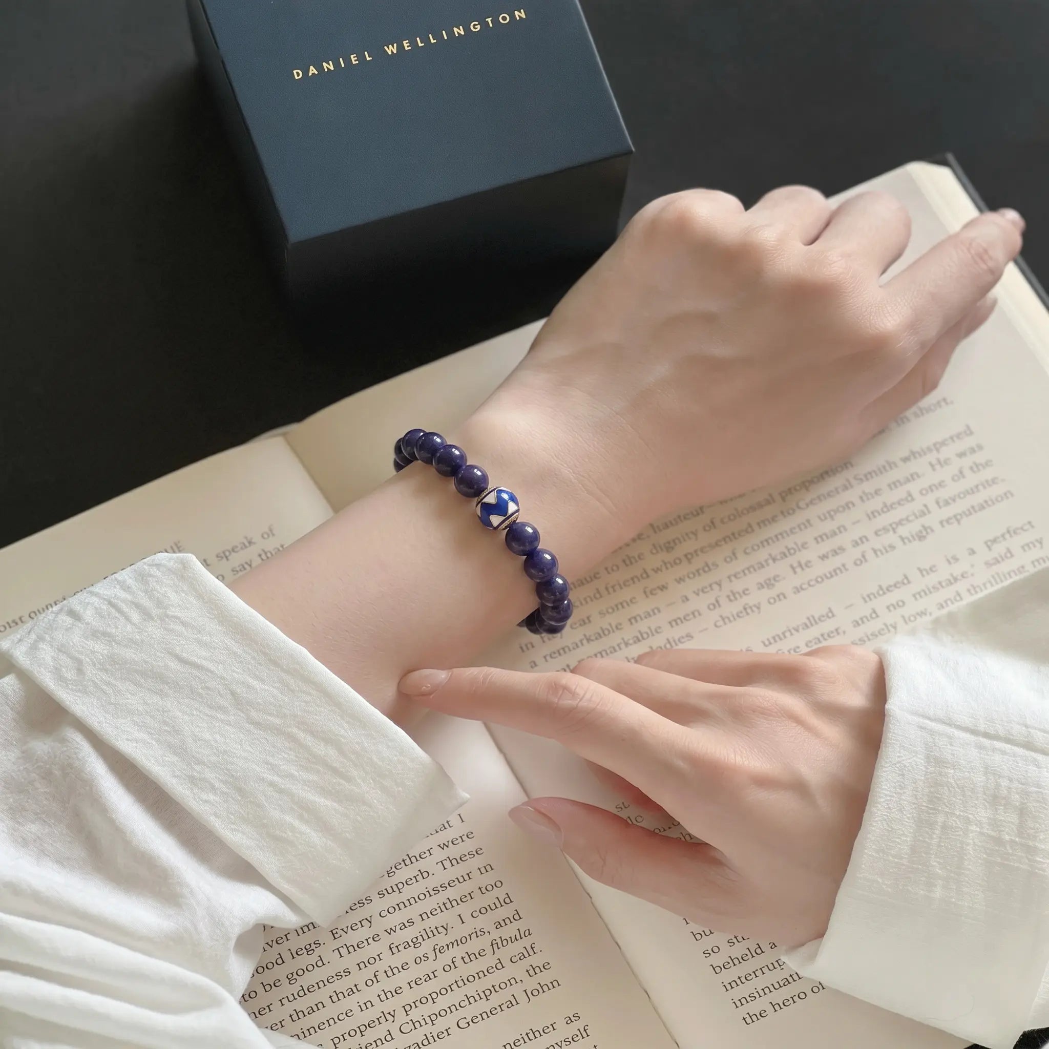 Macro diagonal view of a left hand resting flat on an open book while right-hand fingers touch the lapis lazuli wrist bracelet.