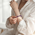 Right-side perspective close-up of a lady with her left elbow resting on a desk and raised forearm showing an amethyst bracelet touched by her right hand.