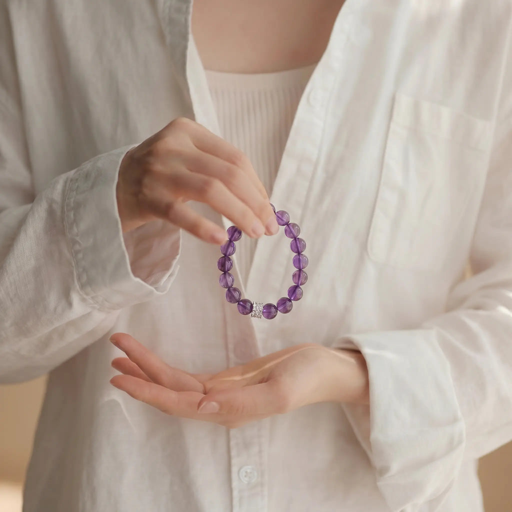 Cropped upper body shot to the collarbone showing a spiritual cupping gesture with hands holding a purple crystal bracelet near the chest.