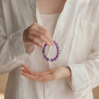 Cropped upper body shot to the collarbone showing a spiritual cupping gesture with hands holding a purple crystal bracelet near the chest.