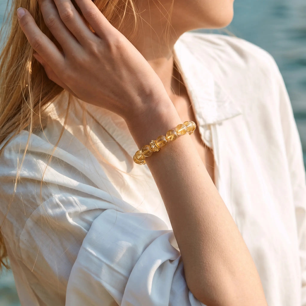 Low-angle profile perspective of an elegant woman touching her hair with a raised hand revealing a golden gemstone bracelet.