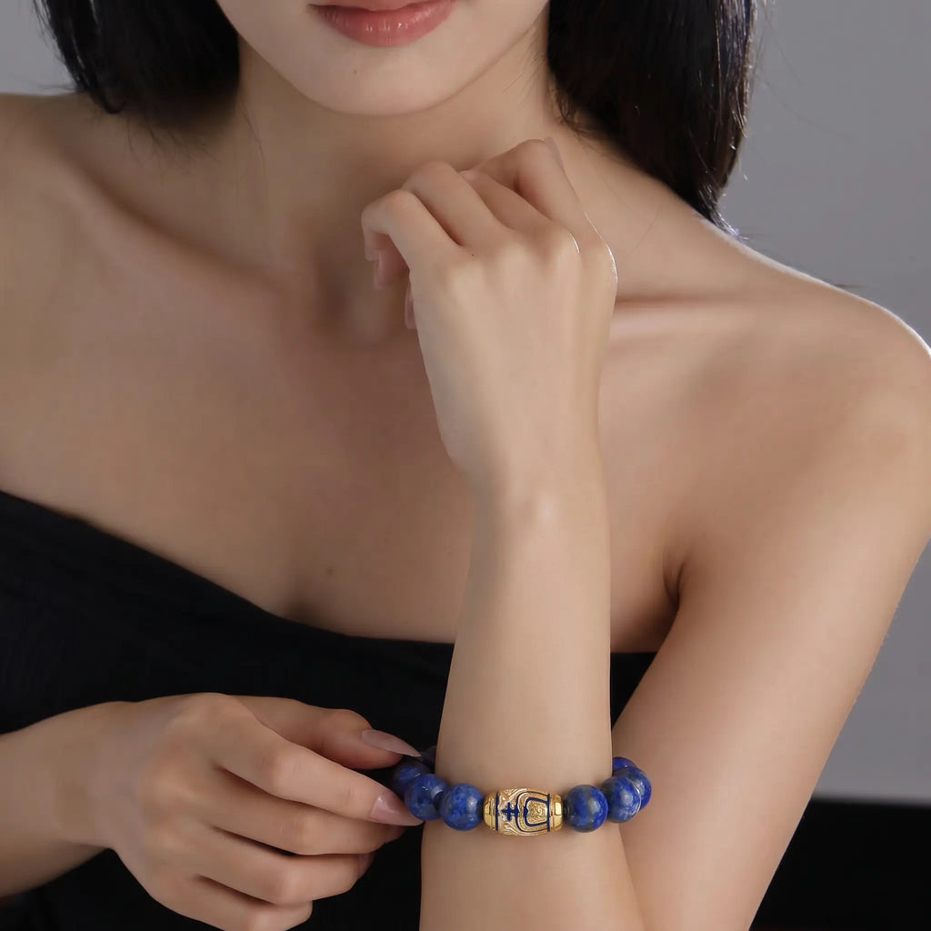 Front close-up view from collarbone to chest showing a woman right wrist wearing the Tranquil Lapis Bracelet with left fingers gently touching the centerpiece.