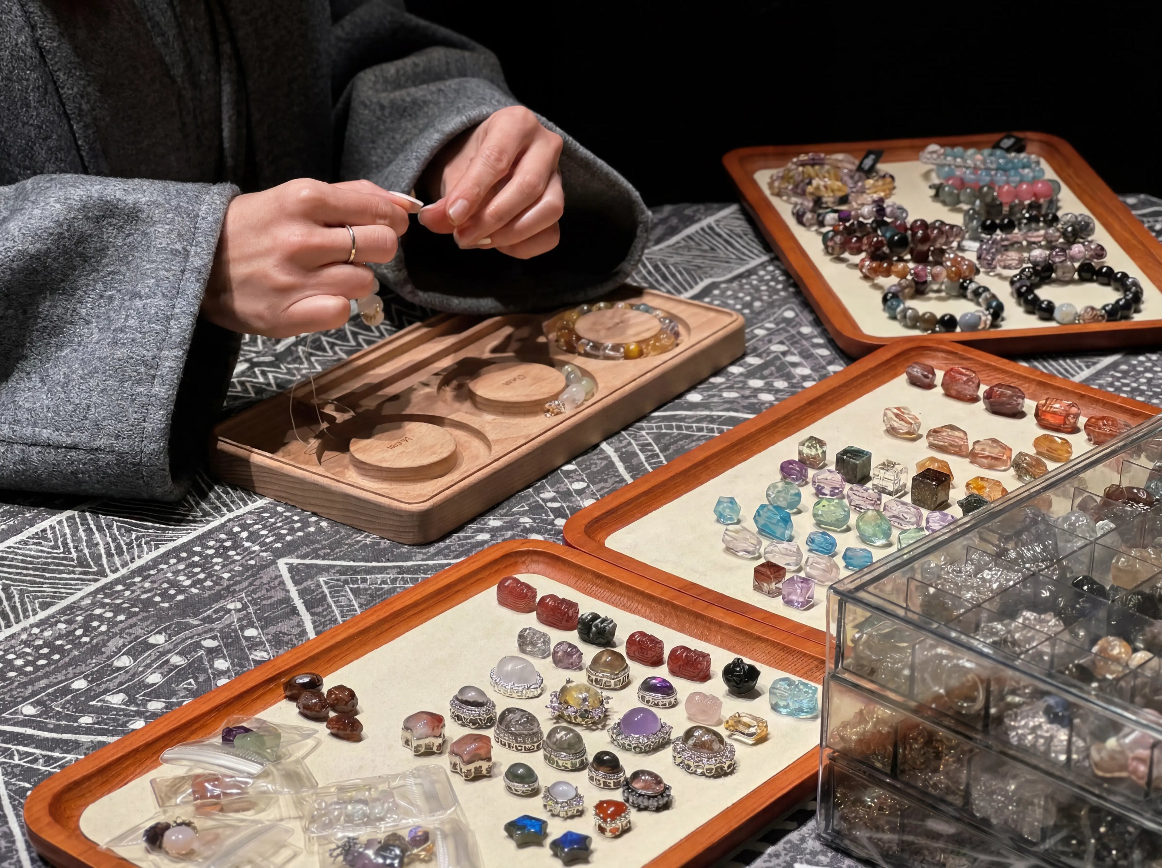 Front 45 degree angle view of a female artisan hand-stringing gemstone beads at a workshop table full of jewelry supplies.