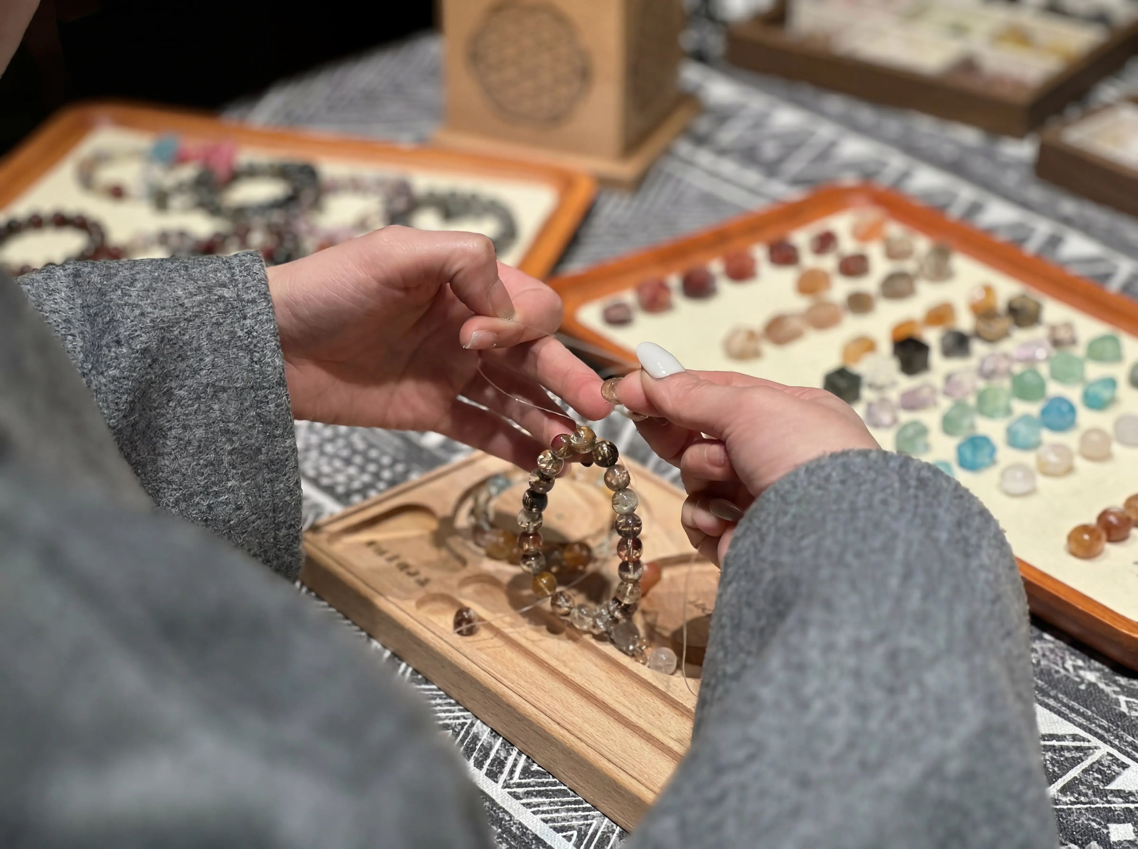 Back 45 degree angle view of a female jewelry maker stringing crystal beads at a workspace with diverse beads and tools.